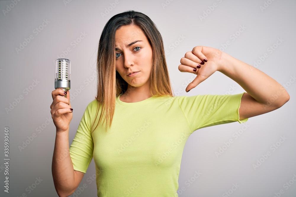 Young beautiful blonde woman holding led lightbulb over isolated white background with angry face, negative sign showing dislike with thumbs down, rejection concept Young beautiful blonde woman holding led lightbulb over isolated white background with angry face, negative sign showing dislike with thumbs down, rejection concept