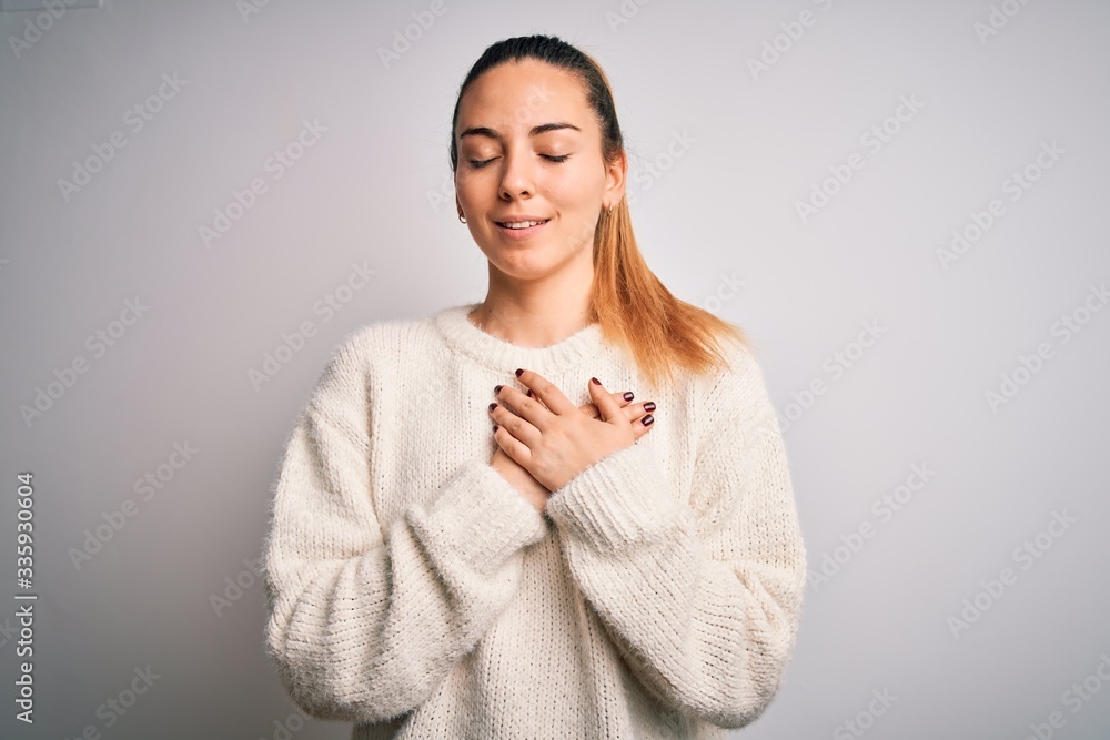 Young beautiful blonde woman with blue eyes wearing casual sweater over white background smiling with hands on chest with closed eyes and grateful gesture on face. Health concept. Young beautiful blonde woman with blue eyes wearing casual sweater over white background smiling with hands on chest with closed eyes and grateful gesture on face. Health concept.