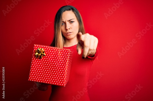 Young beautiful blonde woman with blue eyes holding birthday gift over red background pointing with finger to the camera and to you, hand sign, positive and confident gesture from the front © Krakenimages.com