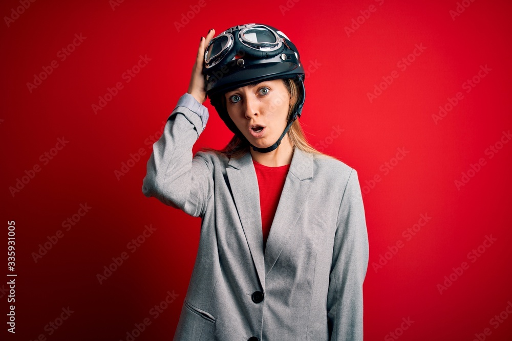 Young beautiful blonde motorcyclist woman wearing motorcycle helmet over red background surprised with hand on head for mistake, remember error. Forgot, bad memory concept. Young beautiful blonde motorcyclist woman wearing motorcycle helmet over red background surprised with hand on head for mistake, remember error. Forgot, bad memory concept.