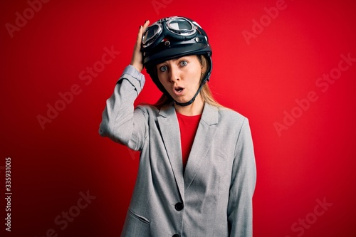 Young beautiful blonde motorcyclist woman wearing motorcycle helmet over red background surprised with hand on head for mistake, remember error. Forgot, bad memory concept.