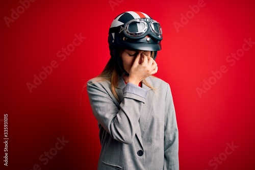 Young beautiful blonde motorcyclist woman wearing motorcycle helmet over red background tired rubbing nose and eyes feeling fatigue and headache. Stress and frustration concept.