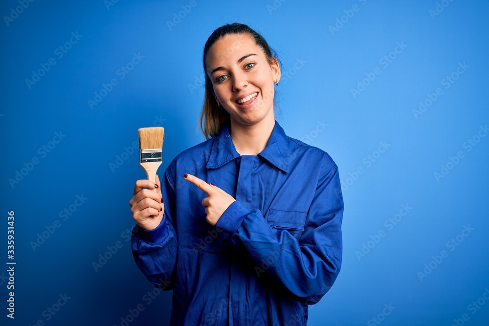 Young beautiful painter woman with blue eyes painting wearing uniform using paint brush very happy pointing with hand and finger Young beautiful painter woman with blue eyes painting wearing uniform using paint brush very happy pointing with hand and finger