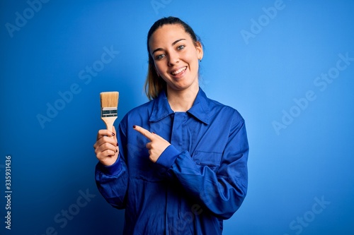 Young beautiful painter woman with blue eyes painting wearing uniform using paint brush very happy pointing with hand and finger