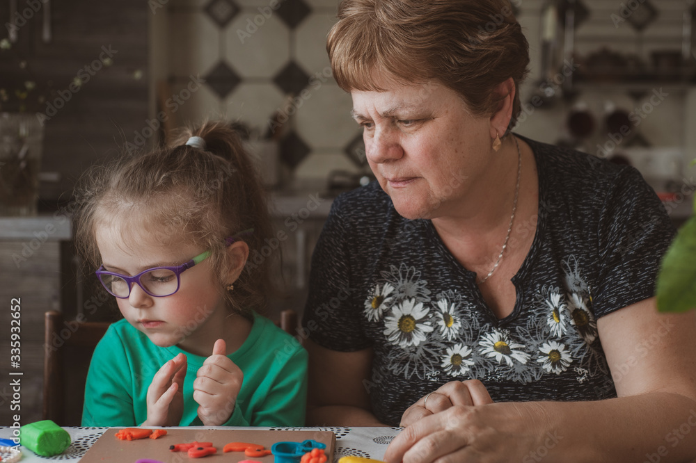 An adult woman and a little girl in glasses make figures from plasticine at home. Grandmother and granddaughter are sitting at home. Family Values Concept An adult woman and a little girl in glasses make figures from plasticine at home. Grandmother and granddaughter are sitting at home. Family Values Concept