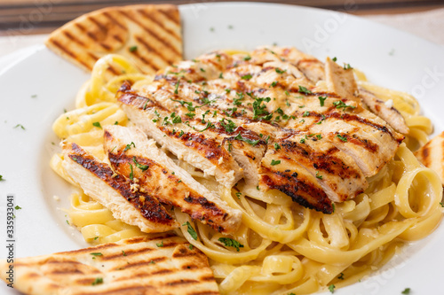 A closeup view of a plate of fettuccine alfredo with grilled chicken breast, in a restaurant or kitchen setting.
