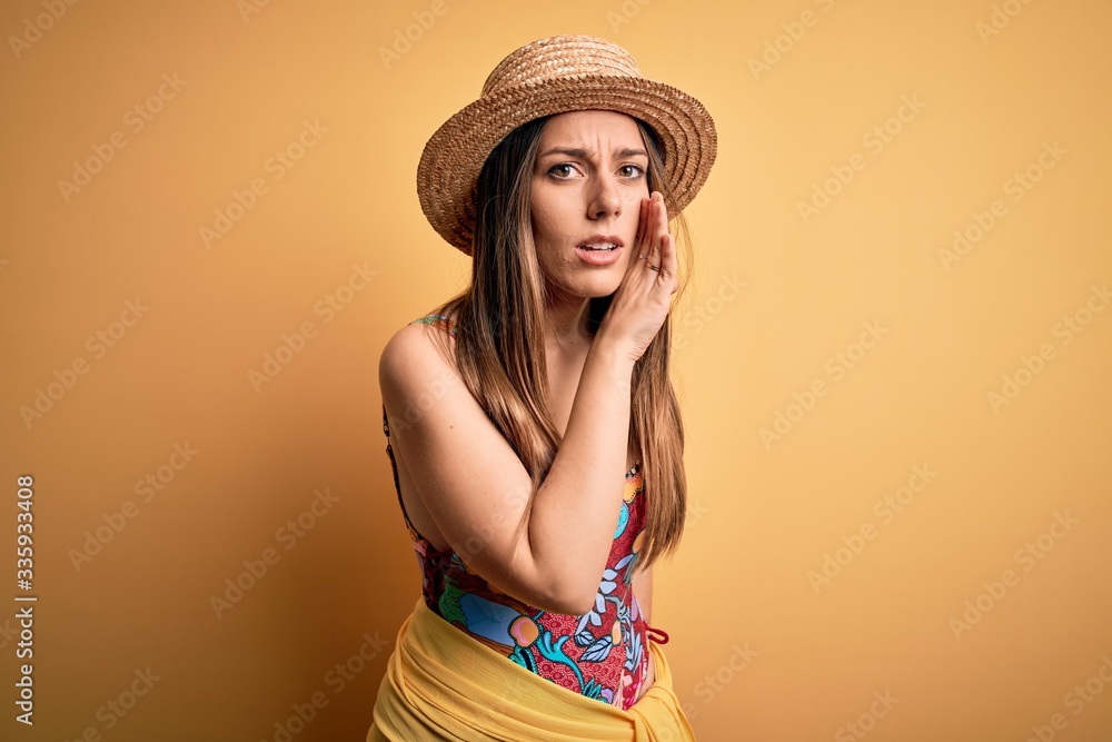 Young beautiful blonde woman wearing swimsuit and summer hat over yellow background hand on mouth telling secret rumor, whispering malicious talk conversation Young beautiful blonde woman wearing swimsuit and summer hat over yellow background hand on mouth telling secret rumor, whispering malicious talk conversation