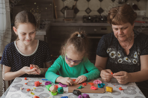 An adult woman and two little girls make plasticine figurines at home. Grandmother and granddaughters are sitting at home. Family Values Concept
