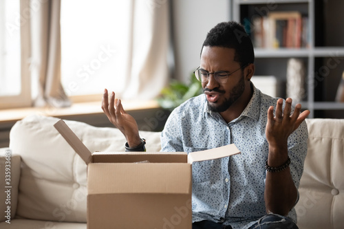 Unhappy African American man client disappointed with product quality shopping online, mad biracial male open cardboard package frustrated with wrong Internet order, bad delivery service concept photo