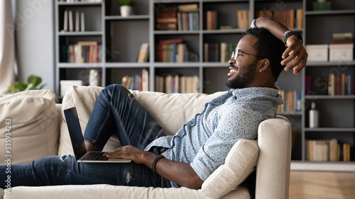Happy biracial young man relax on comfortable sofa at home working on modern computer gadget, smiling African American millennial male rest on cozy couch in living room using laptop device