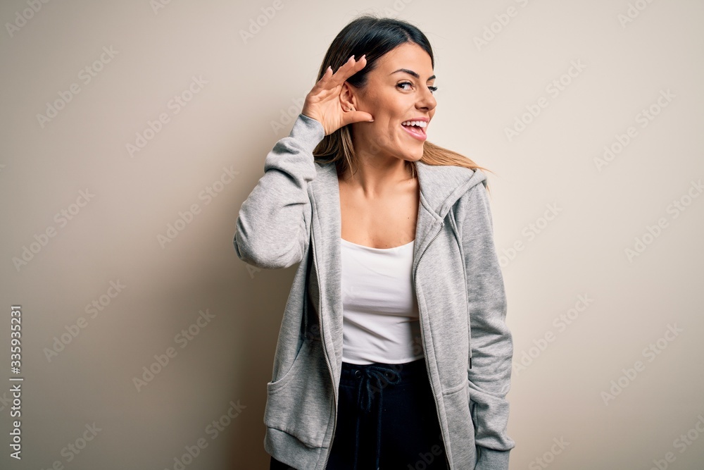 Young beautiful brunette sportswoman wearing sportswoman training over white background smiling with hand over ear listening an hearing to rumor or gossip. Deafness concept. Young beautiful brunette sportswoman wearing sportswoman training over white background smiling with hand over ear listening an hearing to rumor or gossip. Deafness concept.