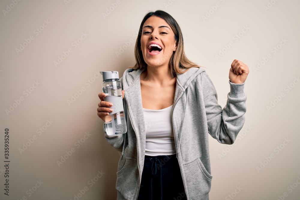 Young beautiful brunette sporty woman drinking bottle of water over isolated white background screaming proud and celebrating victory and success very excited, cheering emotion Young beautiful brunette sporty woman drinking bottle of water over isolated white background screaming proud and celebrating victory and success very excited, cheering emotion