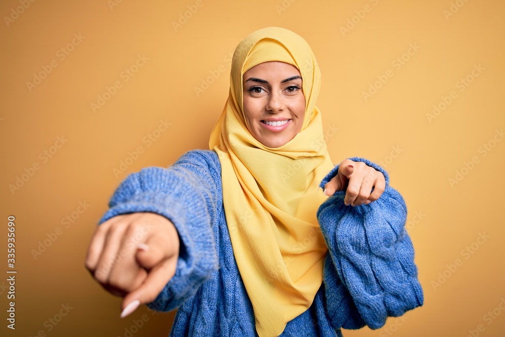 Young beautiful brunette muslim woman wearing arab hijab over isolated yellow background pointing to you and the camera with fingers, smiling positive and cheerful Young beautiful brunette muslim woman wearing arab hijab over isolated yellow background pointing to you and the camera with fingers, smiling positive and cheerful