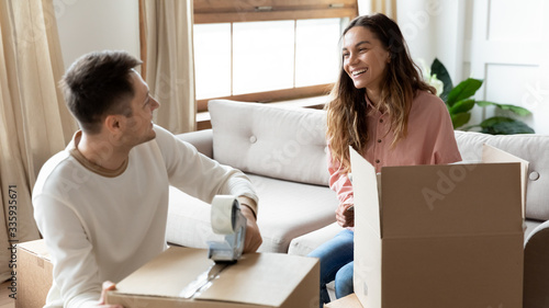 Happy mixed race woman talking with smiling husband while packing home stuff into carton boxes for moving in new house. Joyful family couple preparing belongings for delivery, relocation service.