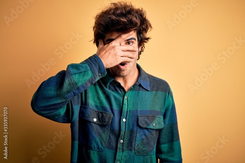 Young handsome man wearing casual shirt standing over isolated yellow background peeking in shock covering face and eyes with hand, looking through fingers with embarrassed expression. © Krakenimages.com