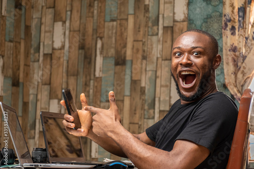 young black man working with his laptop and mobile phone feeling happy and excited pointing to his phone photo