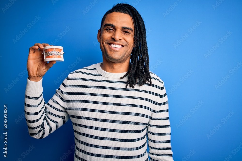 Young african american afro man holding plastic teeth with dental braces with a happy face standing and smiling with a confident smile showing teeth Young african american afro man holding plastic teeth with dental braces with a happy face standing and smiling with a confident smile showing teeth