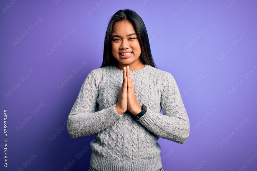 Young beautiful asian girl wearing casual sweater standing over isolated purple background praying with hands together asking for forgiveness smiling confident. Young beautiful asian girl wearing casual sweater standing over isolated purple background praying with hands together asking for forgiveness smiling confident.