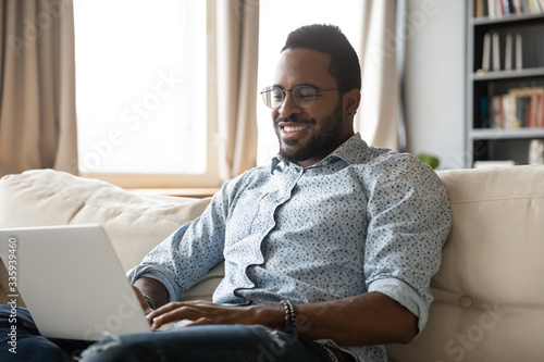Smiling African American millennial man freelancer sit on couch in living room working on modern laptop gadget, happy biracial young male relax on sofa at home browsing wireless Internet on computer
