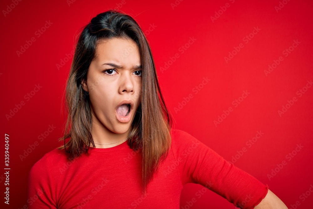 Young beautiful brunette girl wearing casual t-shirt over isolated red background In shock face, looking skeptical and sarcastic, surprised with open mouth Young beautiful brunette girl wearing casual t-shirt over isolated red background In shock face, looking skeptical and sarcastic, surprised with open mouth