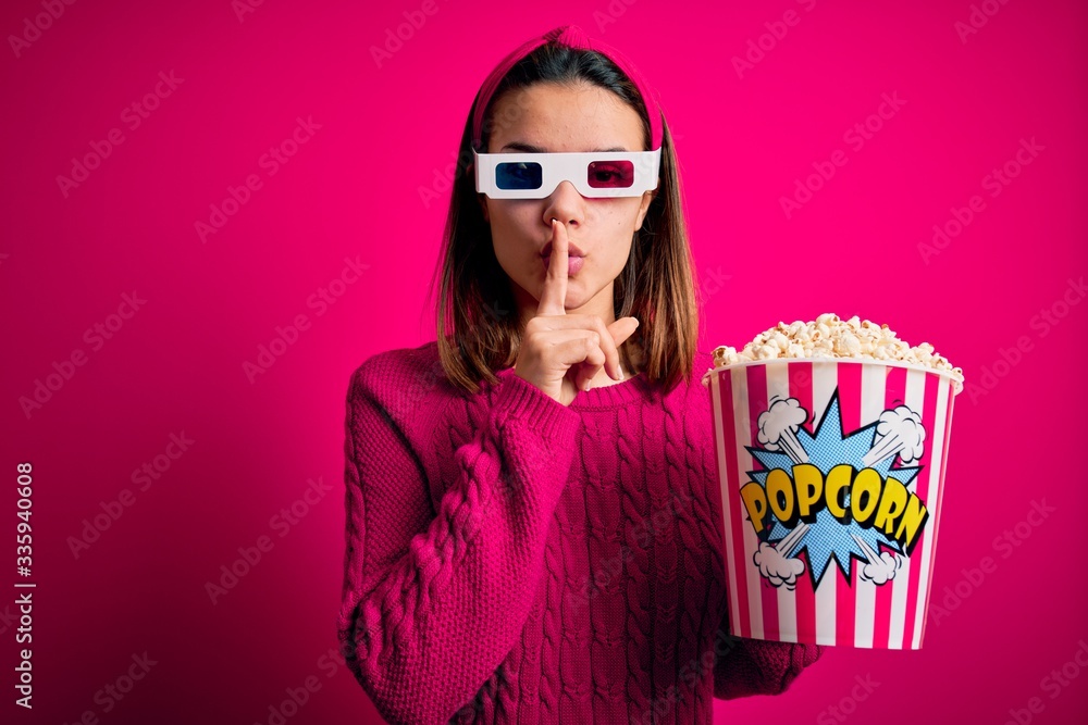 Obraz premium Young beautiful girl watching movie using 3d glasses eating box with popcorns asking to be quiet with finger on lips. Silence and secret concept.