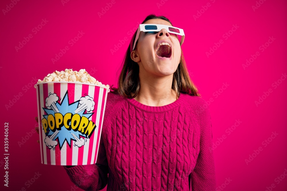 Obraz premium Young beautiful girl watching movie using 3d glasses eating box with popcorns angry and mad screaming frustrated and furious, shouting with anger. Rage and aggressive concept.