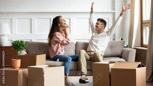 Full length happy married family couple sitting together in living room, laughing celebrating moving in new house. Overjoyed homeowners feeling excited about apartment flat mortgage purchase.