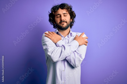 Young handsome business man with beard wearing shirt standing over purple background Hugging oneself happy and positive, smiling confident. Self love and self care © Krakenimages.com
