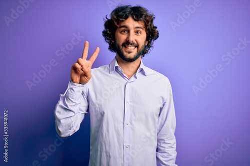 Young handsome business man with beard wearing shirt standing over purple background showing and pointing up with fingers number two while smiling confident and happy.