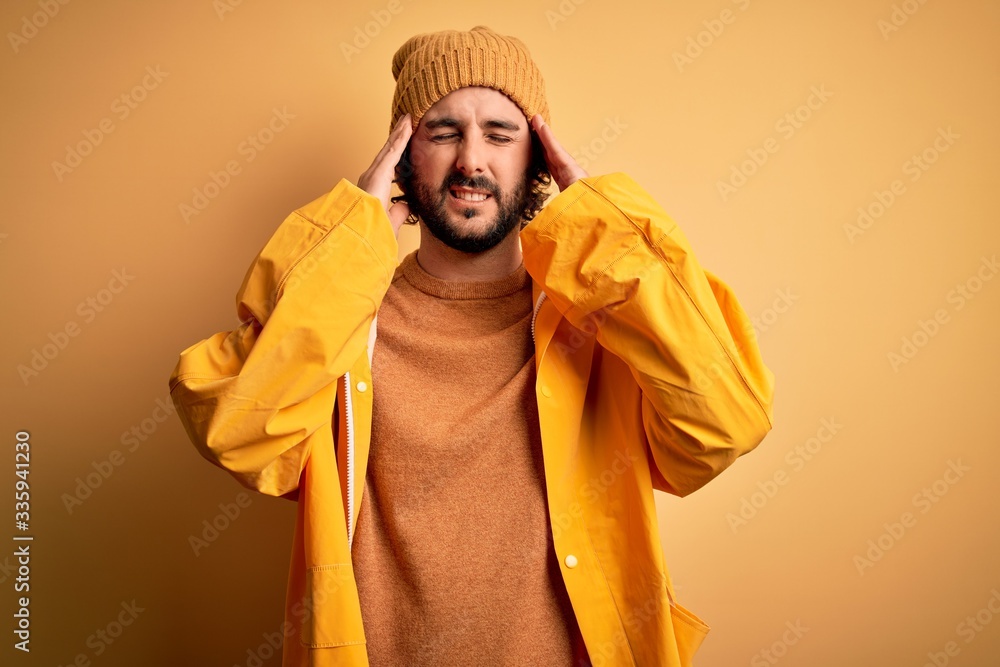 Young handsome man with beard wearing raincoat for rainy day over yellow background suffering from headache desperate and stressed because pain and migraine. Hands on head.