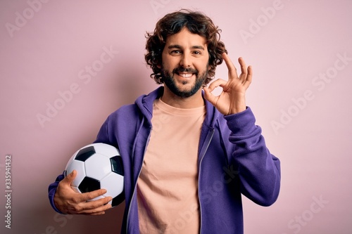 Handsome player man with beard playing soccer holding football ball over pink background doing ok sign with fingers, excellent symbol