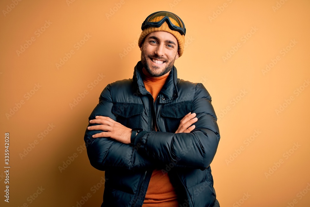 Young handsome skier man with beard wearing snow sportswear and ski goggles happy face smiling with crossed arms looking at the camera. Positive person. Young handsome skier man with beard wearing snow sportswear and ski goggles happy face smiling with crossed arms looking at the camera. Positive person.