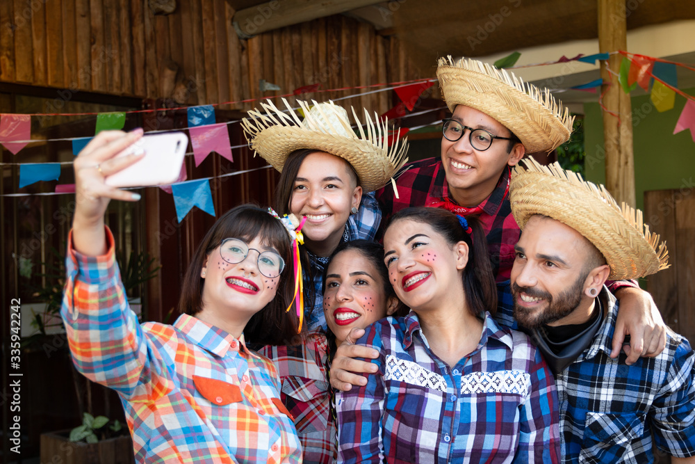 Festa Junina in Brazil, known as brazilian June Party. Group of people in  traditional plaid clothes taking a selfie, looking at cell phone screen.  Rustic wooden decor with flags.. Stock Photo