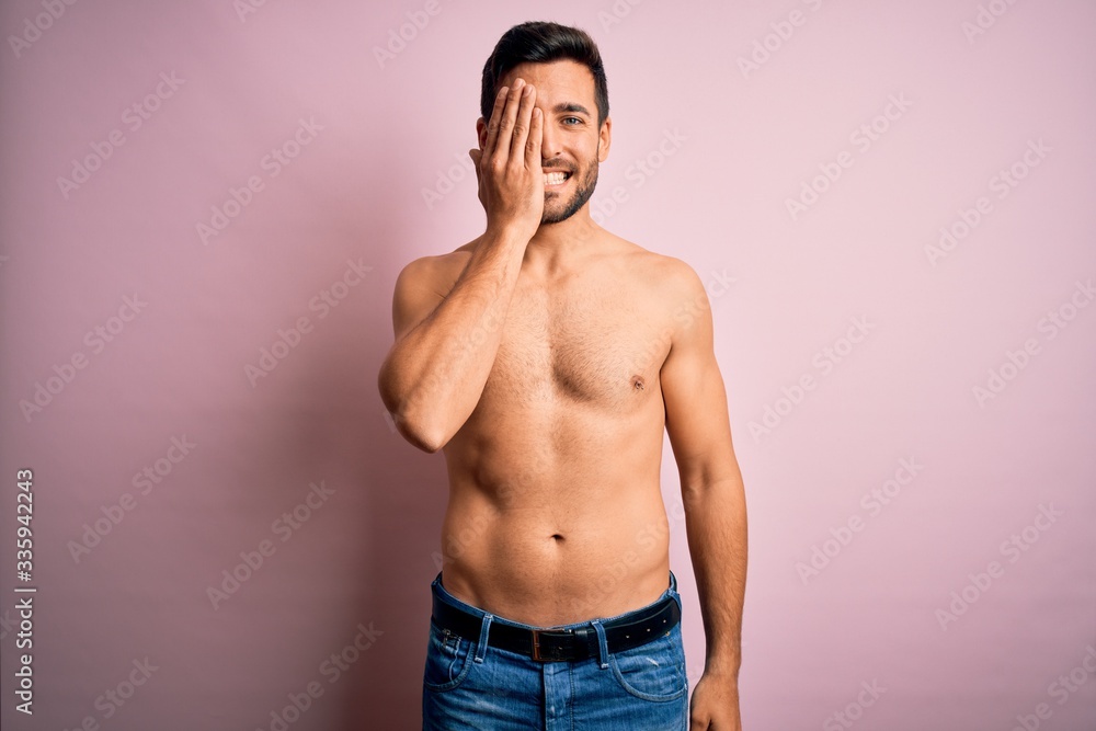 Young handsome strong man with beard shirtless standing over isolated pink background covering one eye with hand, confident smile on face and surprise emotion.