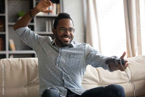Happy African American young man in glasses sit on couch at home play video game, overjoyed biracial millennial male gamer relax in living room engaged in digital virtual activity
