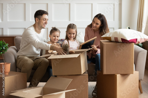 Happy couple sitting on couch, unboxing family belongings with adorable two daughters in new house. Smiling parents unpacking cardboard boxes with toys and decoration with small children t home.