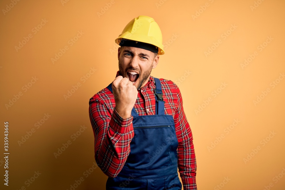 Young builder man wearing construction uniform and safety helmet over yellow isolated background angry and mad raising fist frustrated and furious while shouting with anger. Rage and aggressive 