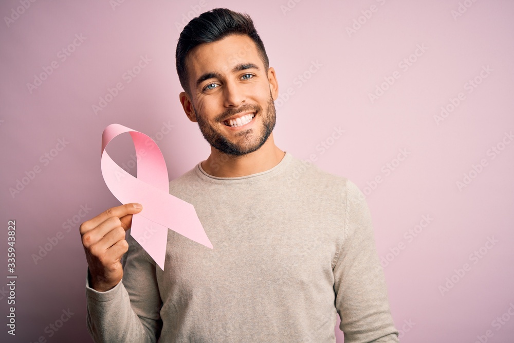 Young handsome man holding pink cancer ribbon symbol of support over isolated background with a happy face standing and smiling with a confident smile showing teeth Young handsome man holding pink cancer ribbon symbol of support over isolated background with a happy face standing and smiling with a confident smile showing teeth