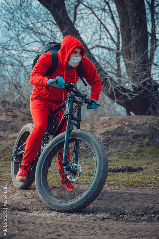 Fototapeta premium Young man in medical mask and gloves, ridding on bicycle in countryside. Male in red suit protecting yourself from diseases on walk. Concept of threat of coronavirus epidemic infection.