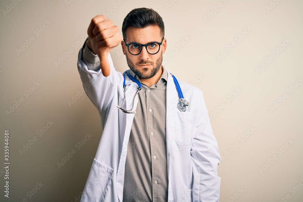Young doctor man wearing glasses, medical white robe and stethoscope over isolated background looking unhappy and angry showing rejection and negative with thumbs down gesture. Bad expression. Young doctor man wearing glasses, medical white robe and stethoscope over isolated background looking unhappy and angry showing rejection and negative with thumbs down gesture. Bad expression.