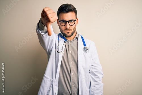Young doctor man wearing glasses, medical white robe and stethoscope over isolated background looking unhappy and angry showing rejection and negative with thumbs down gesture. Bad expression.