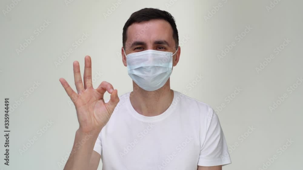 Portrait of joyful male in medical mask is showing OK gesture, looking at the camera, close-up of face. Concept of Coronavirus COVID-19 Pandemic. Studio shot on isolated white background. Portrait of joyful male in medical mask is showing OK gesture, looking at the camera, close-up of face. Concept of Coronavirus COVID-19 Pandemic. Studio shot on isolated white background.