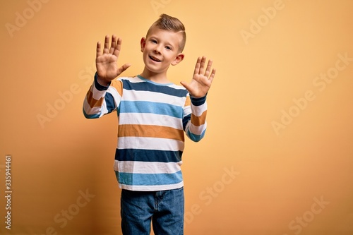 Young little caucasian kid with blue eyes wearing colorful striped shirt over yellow background showing and pointing up with fingers number ten while smiling confident and happy.