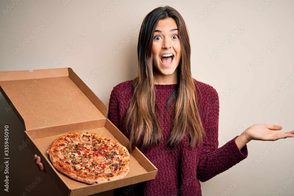 Young beautiful girl holding delivery box with Italian pizza standing over white background very happy and excited, winner expression celebrating victory screaming with big smile and raised hands