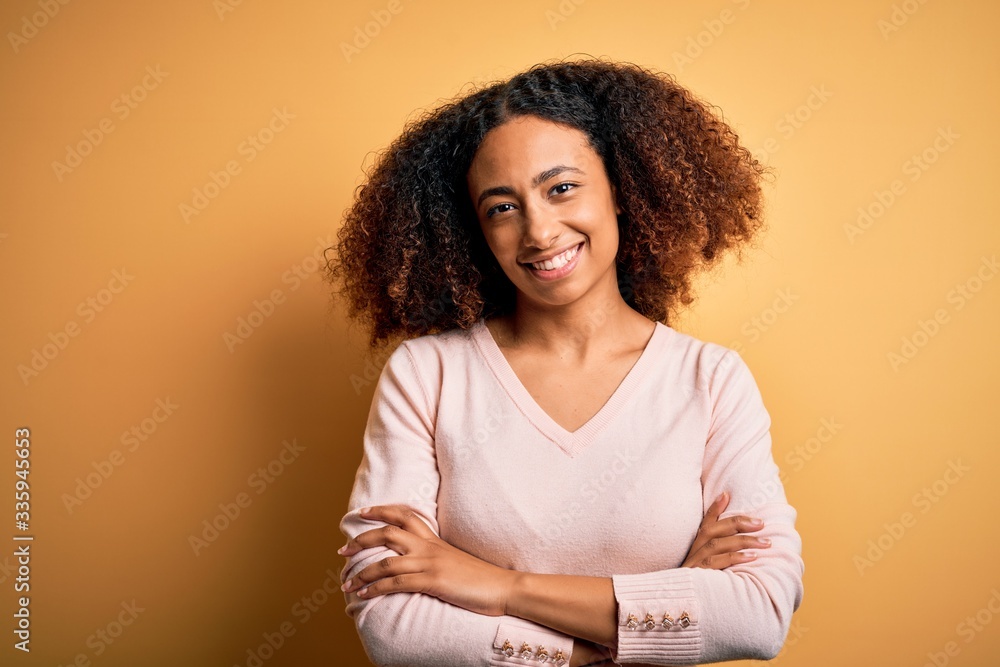 Young african american woman with afro hair wearing casual sweater over yellow background happy face smiling with crossed arms looking at the camera. Positive person. Young african american woman with afro hair wearing casual sweater over yellow background happy face smiling with crossed arms looking at the camera. Positive person.