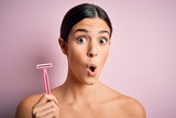 Young beautiful girl using shaver for depilation standing over isolated pink background scared in shock with a surprise face, afraid and excited with fear expression