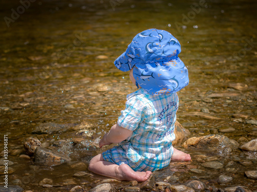 Fototapeta Naklejka Na Ścianę i Meble -  infant boy sits in rocky pond wearing sun protective hat to play