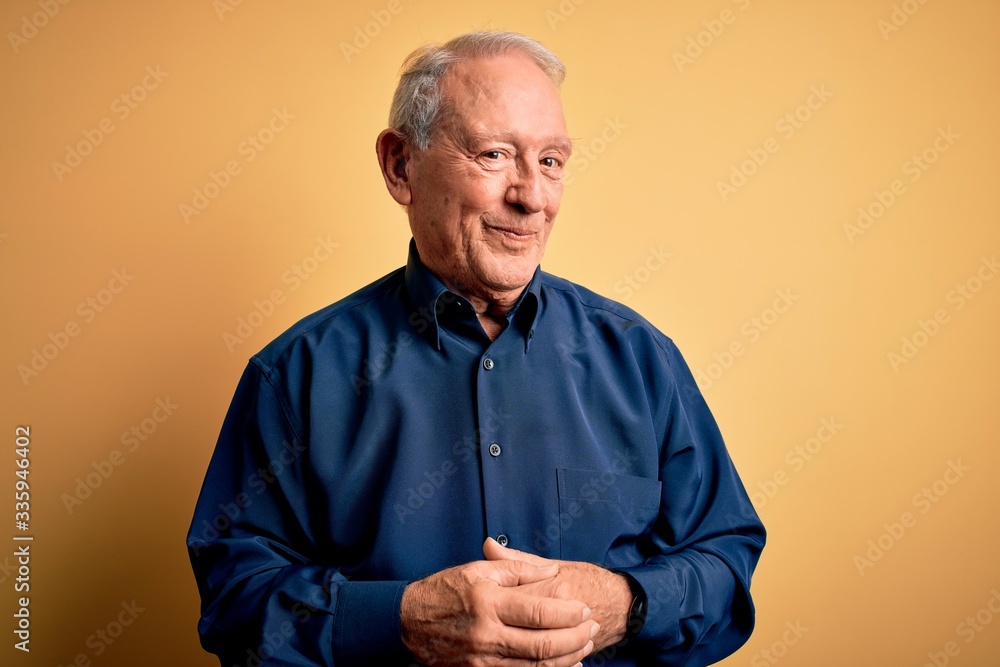 Grey haired senior man wearing casual blue shirt standing over yellow background with hands together and crossed fingers smiling relaxed and cheerful. Success and optimistic Grey haired senior man wearing casual blue shirt standing over yellow background with hands together and crossed fingers smiling relaxed and cheerful. Success and optimistic