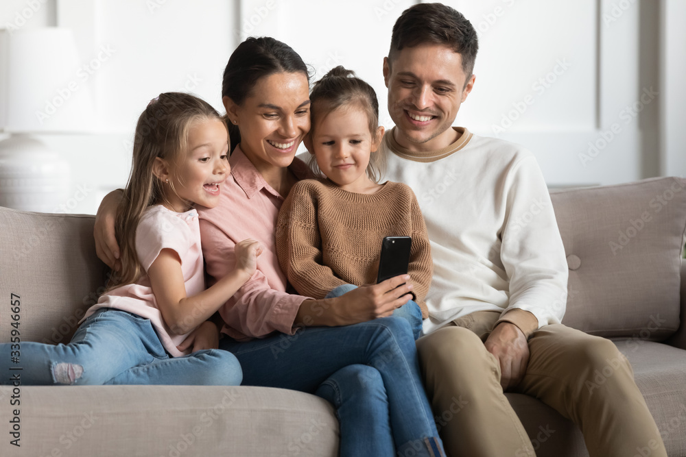 Happy couple relaxing on cozy sofa with cute daughters, looking at mobile phone screen. Joyful family of four spending free weekend time, watching funny video photo on smartphone in living room. Happy couple relaxing on cozy sofa with cute daughters, looking at mobile phone screen. Joyful family of four spending free weekend time, watching funny video photo on smartphone in living room.