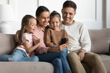 Happy couple relaxing on cozy sofa with cute daughters, looking at mobile phone screen. Joyful family of four spending free weekend time, watching funny video photo on smartphone in living room.